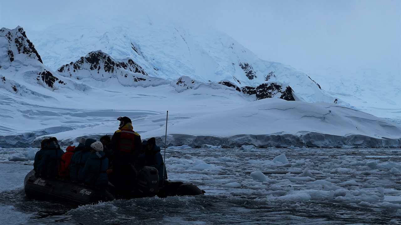 Passengers in cold-weather gear seated in a Zodiac inflatable boat, navigating brash ice against a backdrop of snow-covered mountains on the Antarctic Peninsula